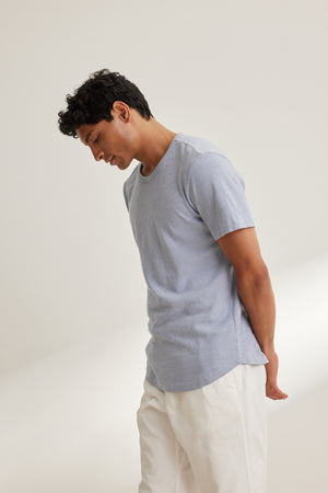 A man wears the SPADER TEE by Velvet by Graham & Spencer—a relaxed fit, light blue Peruvian cotton crewneck—paired with white pants, standing against a plain light background, looking down with hands behind his back.