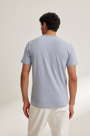 A man faces away from the camera wearing the SPADER TEE by Velvet by Graham & Spencer—a relaxed fit, light blue short-sleeve crew neck t-shirt—with white pants, set against a plain light background.