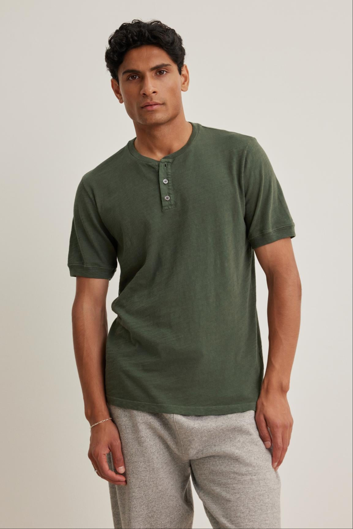 A man confidently stands against a plain backdrop wearing the Velvet by Graham & Spencer VITO HENLEY in green, made from soft Peruvian cotton, paired with gray pants.-38643367477441
