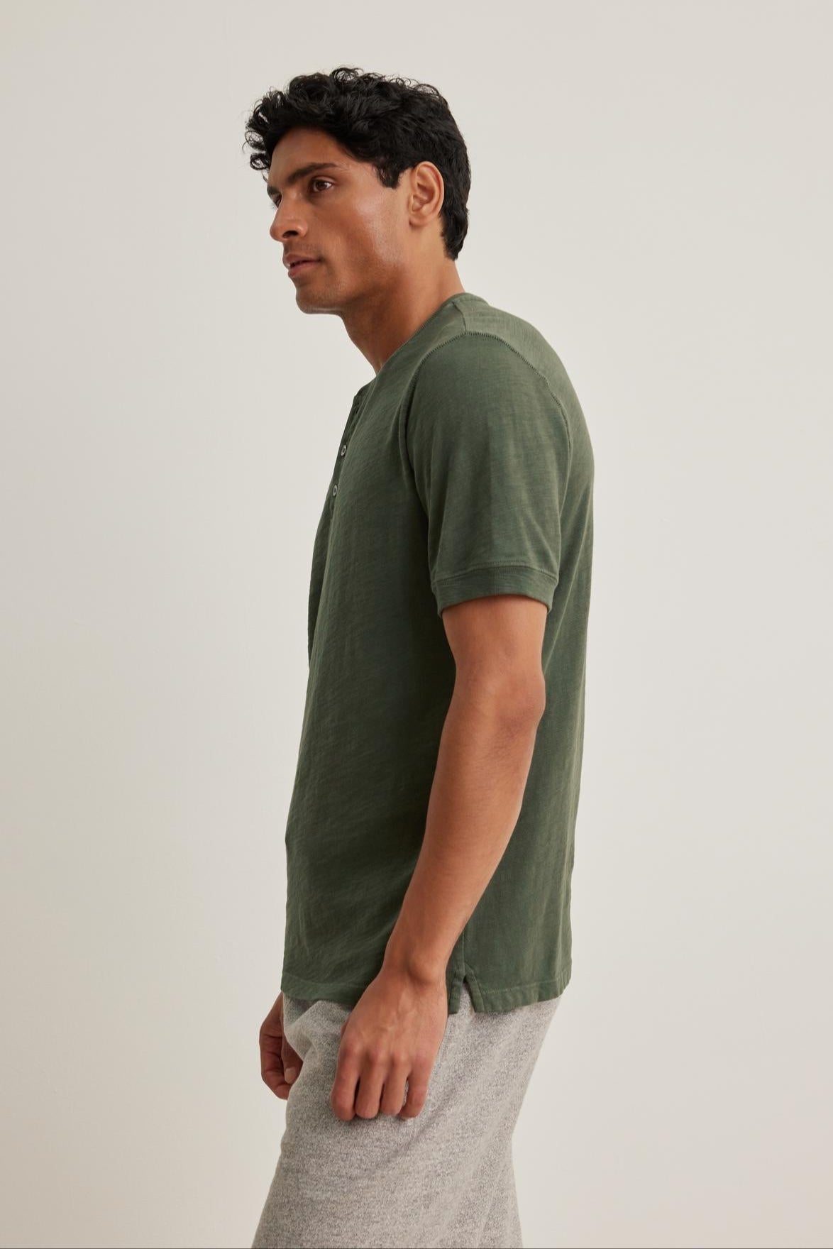 A person in a VITO HENLEY by Velvet by Graham & Spencer, featuring a textured feel and short sleeves in green, paired with gray pants, stands facing left against a simple background.-38643367444673