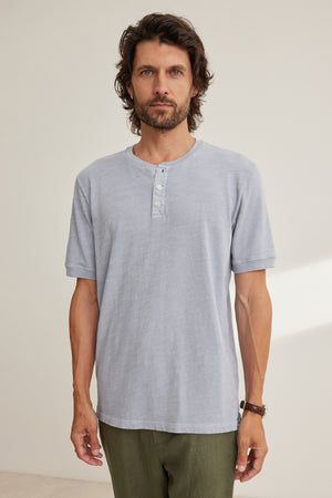 A man with medium-length brown hair and a beard wears the Velvet by Graham & Spencer VITO HENLEY, a light gray Peruvian cotton shirt with a textured feel, paired with olive green pants and standing against a plain background.