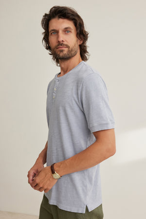Man with wavy brown hair and beard wears the VITO HENLEY by Velvet by Graham & Spencer, a light gray Peruvian cotton short-sleeve shirt with three buttons and a watch, standing against a plain light background.