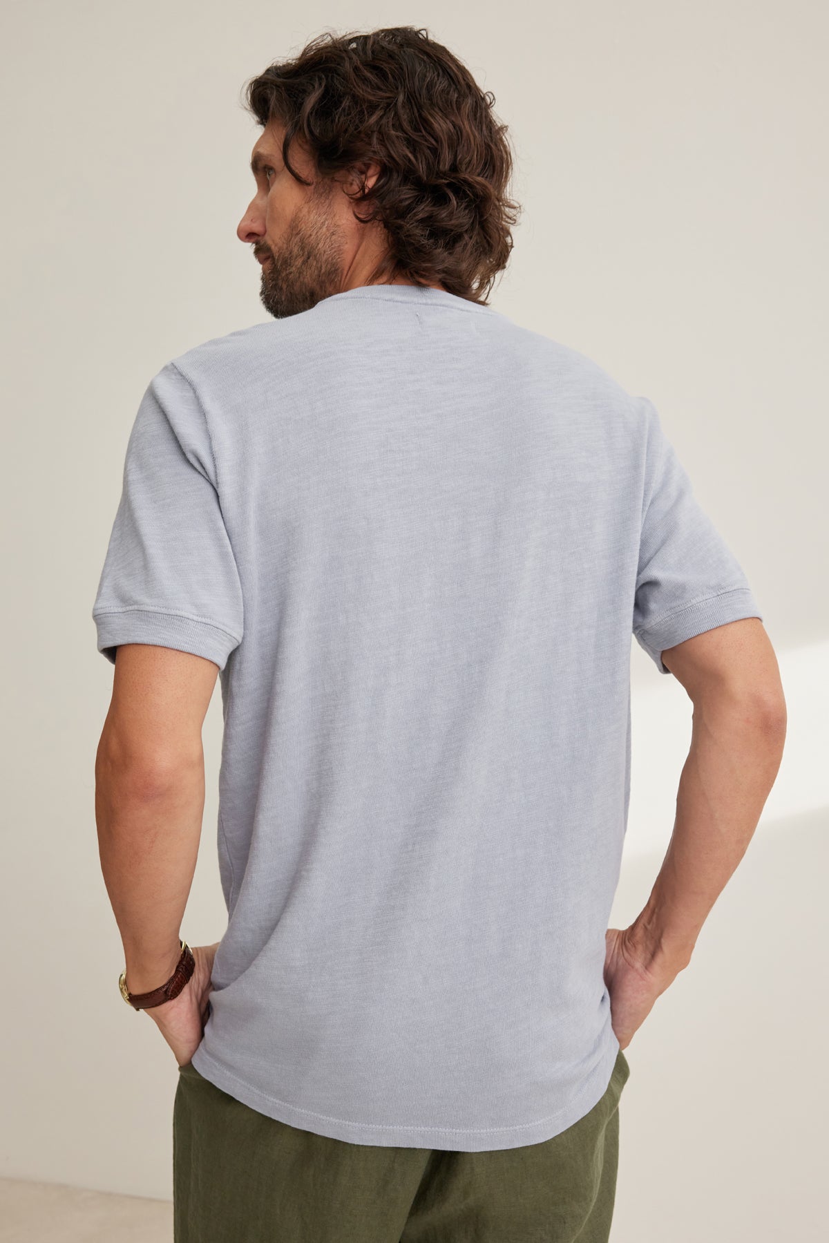 A man with medium-length brown hair stands facing away, wearing a light gray VITO HENLEY by Velvet by Graham & Spencer and olive green pants, his right hand on his hip.-39152759505089
