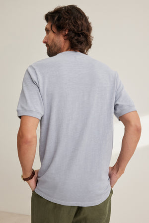 A man with medium-length brown hair stands facing away, wearing a light gray VITO HENLEY by Velvet by Graham & Spencer and olive green pants, his right hand on his hip.