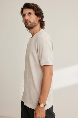A man with shoulder-length hair and beard wears the VITO HENLEY, a light short-sleeve shirt from Velvet by Graham & Spencer made of Peruvian cotton for a textured feel. He stands against a plain backdrop, looking sideways as his watch subtly catches the light.