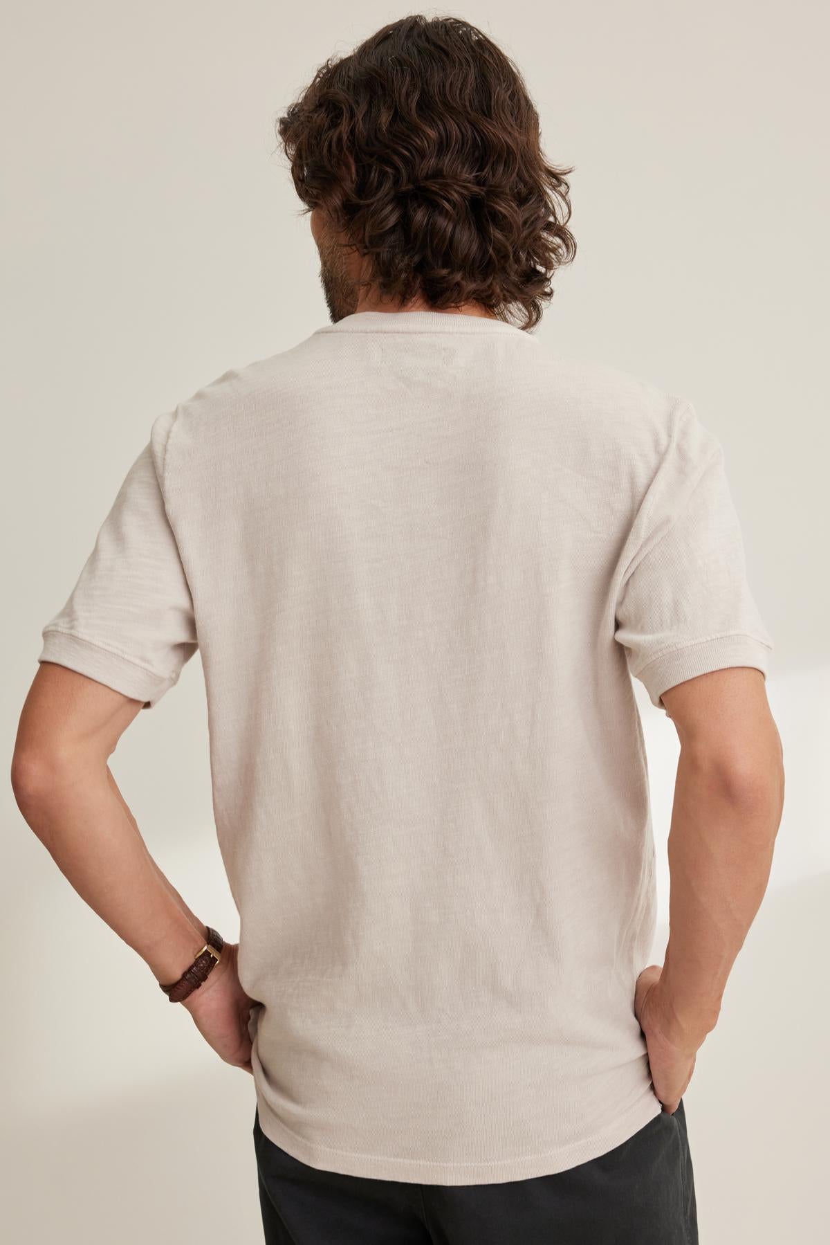 Seen from the back, a person with curly hair stands hands on hips, wearing the VITO HENLEY by Velvet by Graham & Spencer in beige Peruvian cotton with a textured feel and dark pants against a light background.-38643365740737