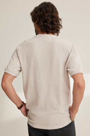 Seen from the back, a person with curly hair stands hands on hips, wearing the VITO HENLEY by Velvet by Graham & Spencer in beige Peruvian cotton with a textured feel and dark pants against a light background.