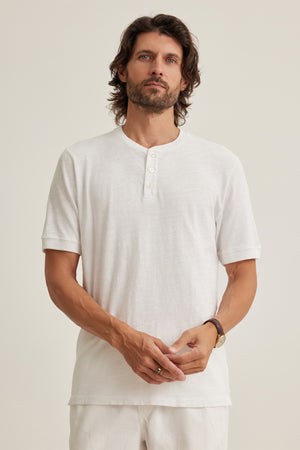 A man with wavy brown hair and a beard wears the Velvet by Graham & Spencer VITO HENLEY, a white short-sleeve Peruvian cotton shirt, with white pants, standing against a plain light background.