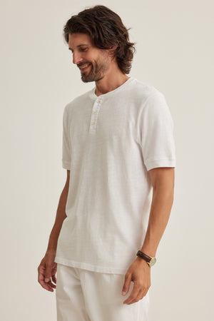 A man with medium-length brown hair and a beard is wearing the VITO HENLEY by Velvet by Graham & Spencer in white, paired with white pants, standing against a plain light background.