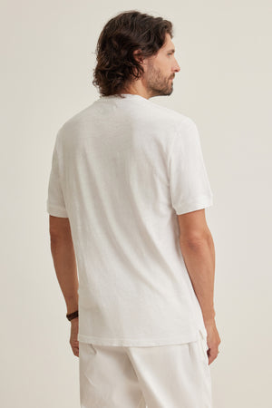 A man with shoulder-length dark hair stands with his back to the camera, wearing the Velvet by Graham & Spencer VITO HENLEY, a plain white Peruvian cotton short-sleeve t-shirt, and white pants against a neutral background.