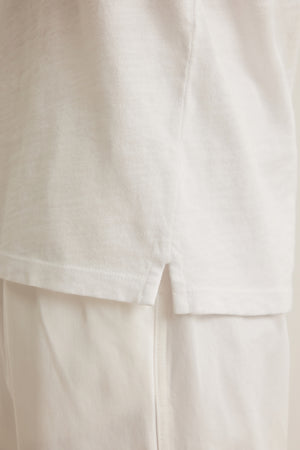 Close-up of the side seam and hem of the VITO HENLEY by Velvet by Graham & Spencer, showing the small slit at the bottom edge and its soft, textured feel in white Peruvian cotton worn over white pants.