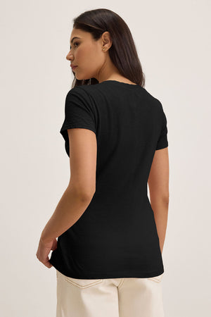 Someone wearing a Velvet by Graham & Spencer LILITH TEE, a luxurious cotton slub V-neck in black, paired with white pants, is facing away from the camera with their head slightly turned to the side, highlighting its flattering fit.