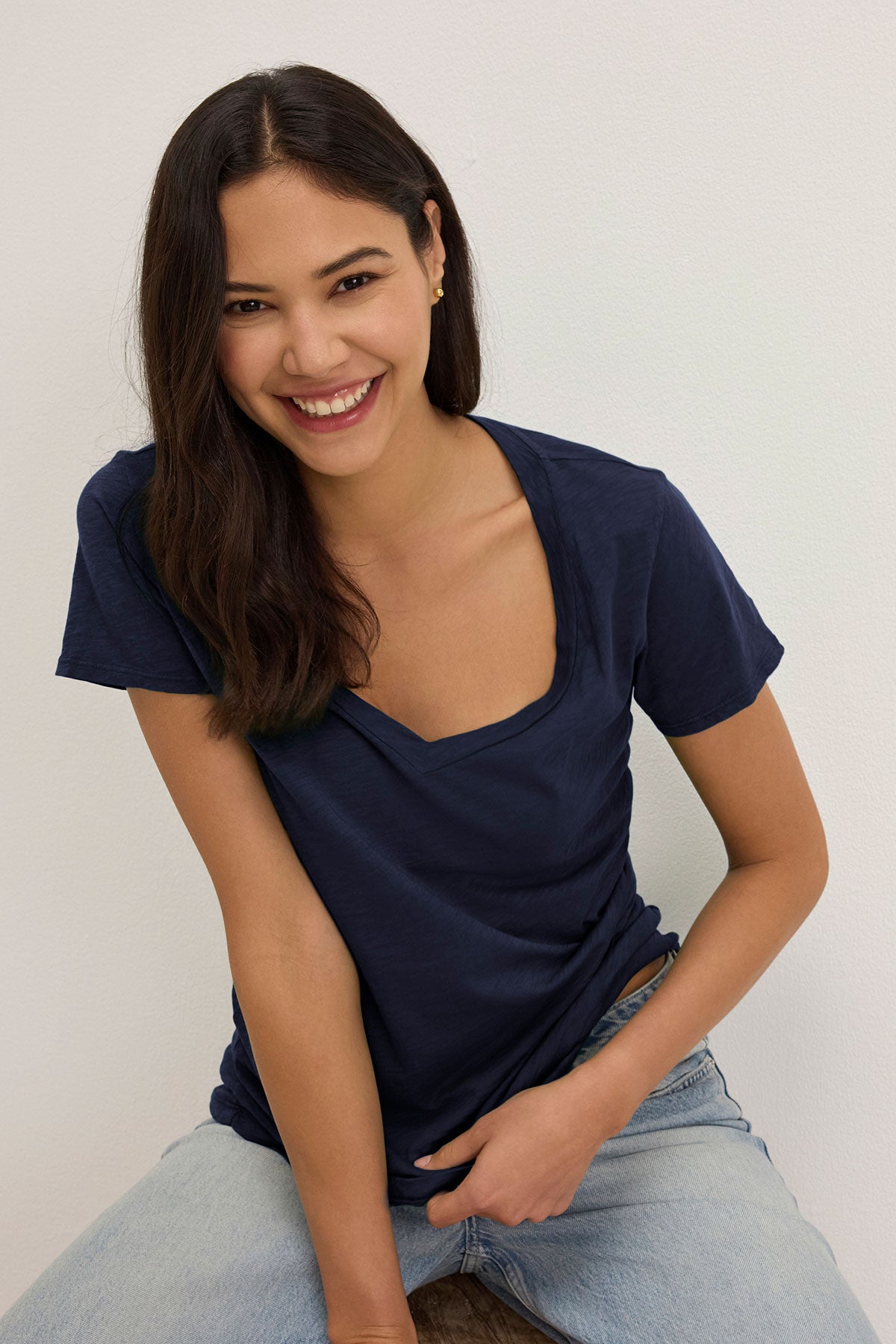 A person with long dark hair, wearing the LILITH TEE by Velvet by Graham & Spencer and jeans, sits and smiles against a plain background. The flattering fit of the cotton slub shirt enhances their relaxed and confident demeanor.-38892104450241