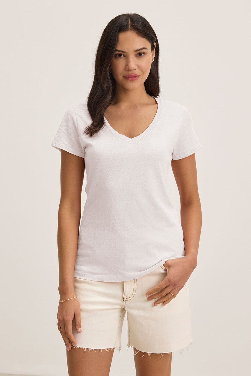 A woman stands against a plain background, wearing the Velvet by Graham & Spencer LILITH TEE—a luxurious cotton slub V-neck tee—paired with light-colored shorts, one hand in her pocket.
