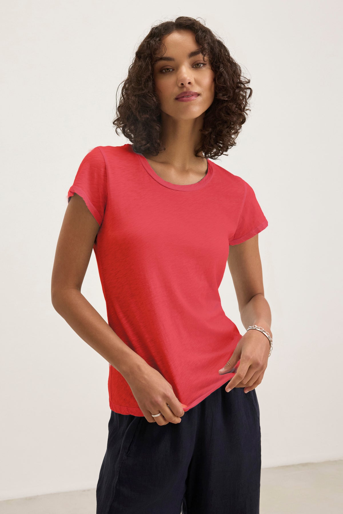 A person in a red ODELIA TEE by Velvet by Graham & Spencer, featuring a classic crew neckline and cozy cotton slub fabric, stands against a plain white background in dark pants, showcasing timeless style and comfort.-38928254861505