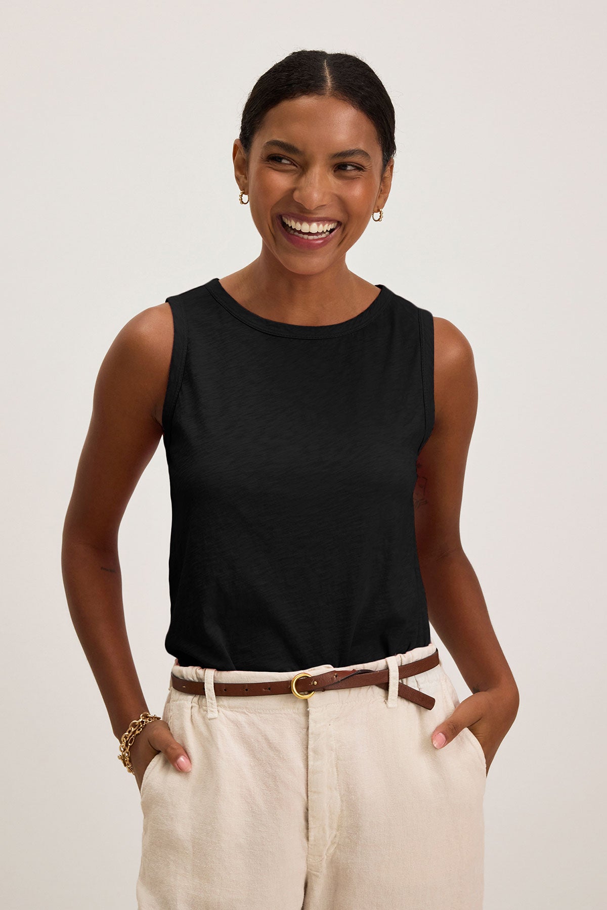 Woman smiling with hands in pockets, wearing the TAURUS TANK TOP by Velvet by Graham & Spencer—a sleeveless black cotton slub top—styled with beige pants, a brown belt, and gold jewelry against a plain light background.-39418761511105