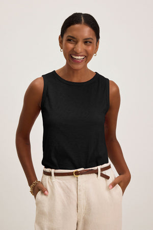 Woman smiling with hands in pockets, wearing the TAURUS TANK TOP by Velvet by Graham & Spencer—a sleeveless black cotton slub top—styled with beige pants, a brown belt, and gold jewelry against a plain light background.