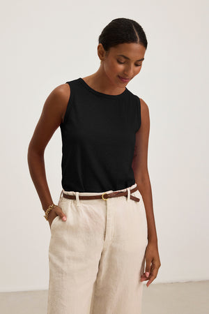 Wearing a Velvet by Graham & Spencer TAURUS TANK TOP and beige pants, a person with short dark hair stands against a plain light background, looking down with hands in pockets.