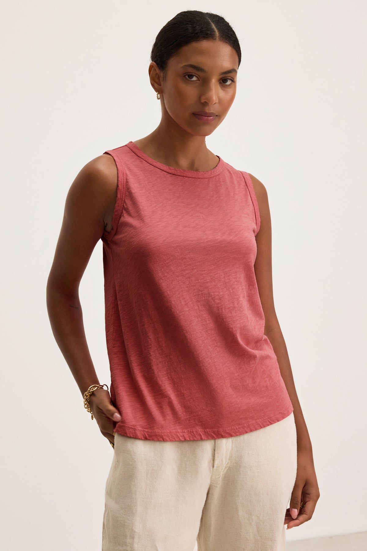 A woman models the TAURUS TANK TOP by Velvet by Graham & Spencer—a sleeveless, rose-colored cotton slub crew neck—paired with light beige pants against a white background, exemplifying effortless style.