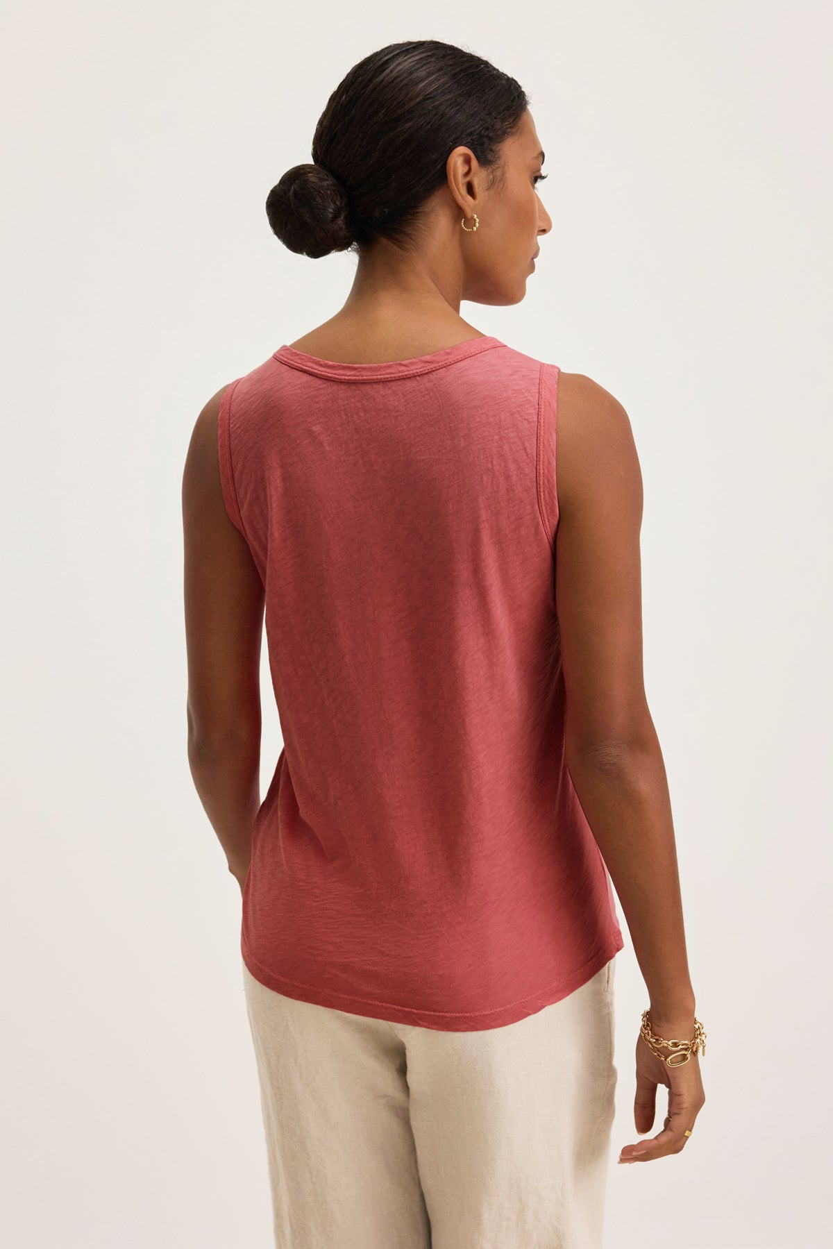 A woman with dark hair in a bun models the Velvet by Graham & Spencer TAURUS TANK TOP, a sleeveless pink textured cotton slub piece, paired with light pants and shown from the back against a plain background.