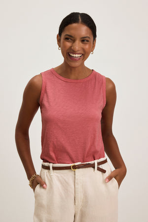 A woman smiles against a plain background, wearing the Velvet by Graham & Spencer TAURUS TANK TOP—a sleeveless rose cotton slub crew neck—styled with beige pants, a brown belt, and gold jewelry.
