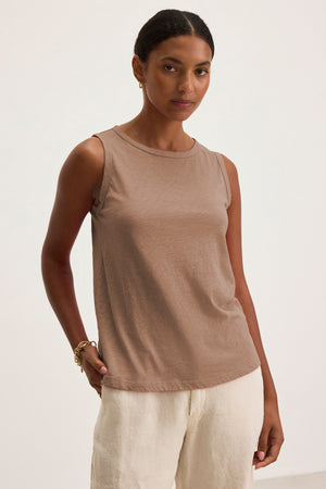 A woman wears the TAURUS TANK TOP by Velvet by Graham & Spencer—a sleeveless taupe crew neck style—paired with light beige pants against a plain background, showcasing an essential piece for effortless style.