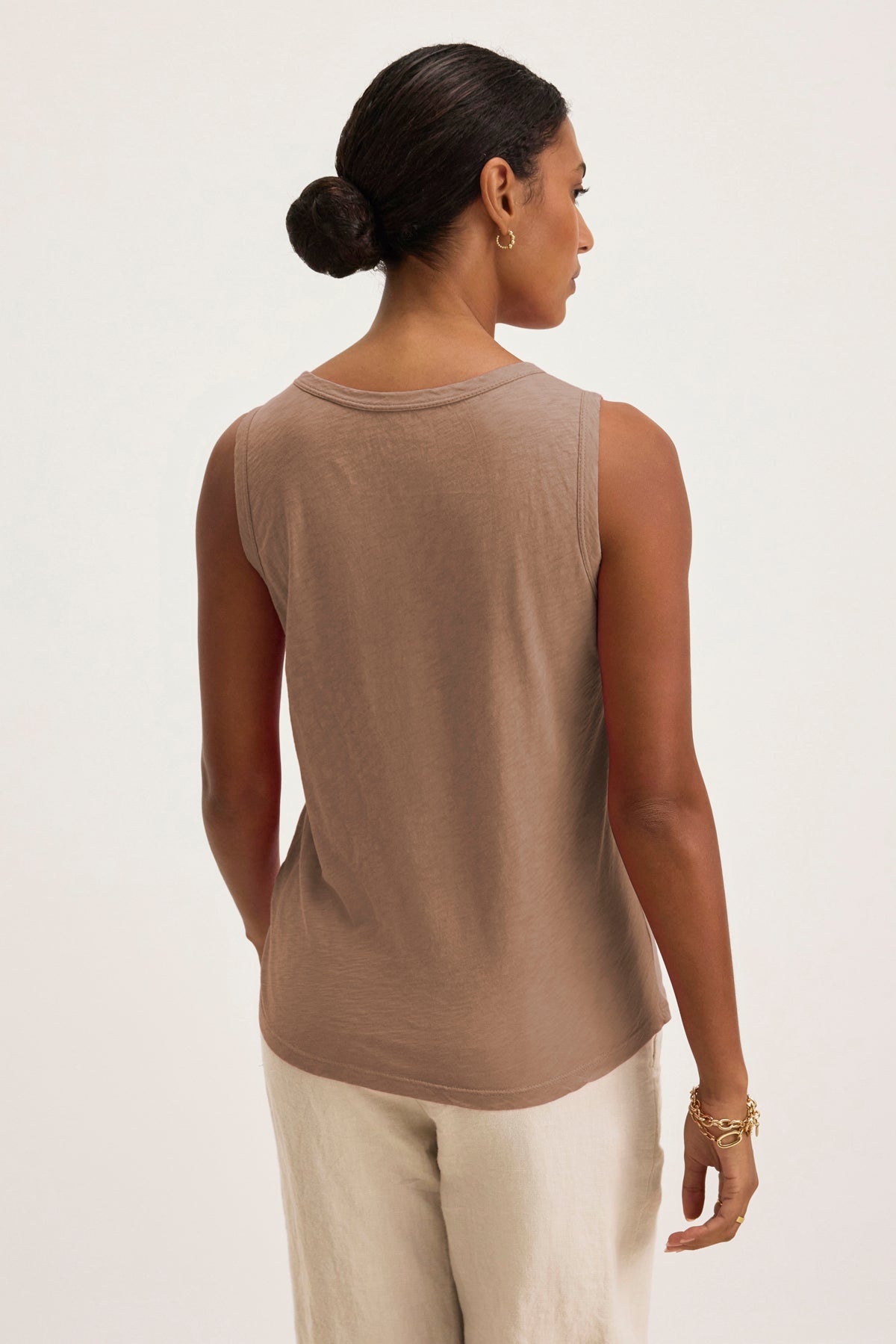 A woman with her hair in a neat bun faces away, wearing the TAURUS TANK TOP by Velvet by Graham & Spencer—a sleeveless brown textured cotton slub top—paired with light-colored pants.