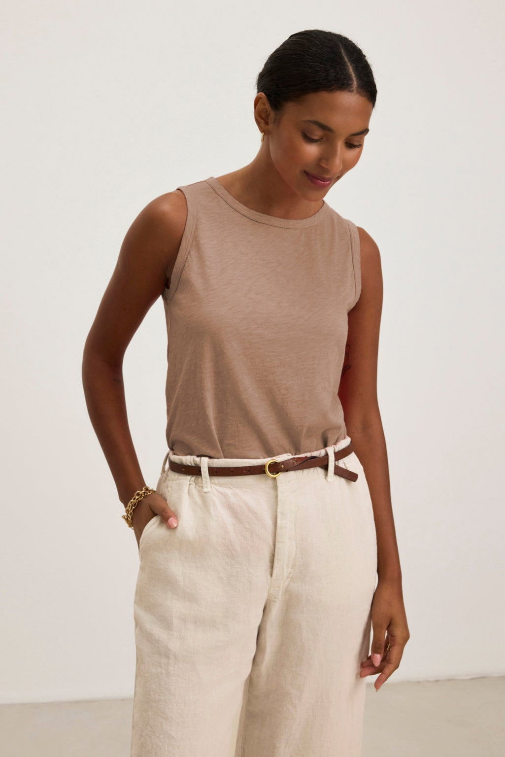 A woman stands against a light background, wearing the TAURUS TANK TOP by Velvet by Graham & Spencer—a sleeveless beige textured cotton slub top with a crew neck—paired with white high-waisted pants, a brown belt, and a gold bracelet, hands in pockets.