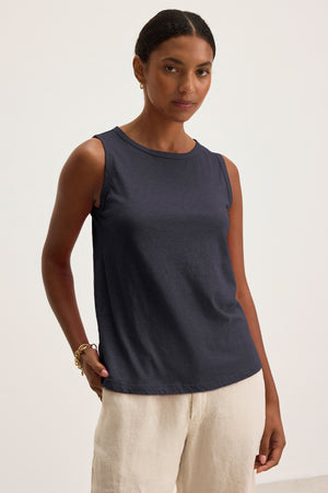 A woman stands against a plain background wearing the TAURUS TANK TOP by Velvet by Graham & Spencer— a navy, textured cotton slub sleeveless top—paired with light-colored linen pants, her left hand in her pocket.