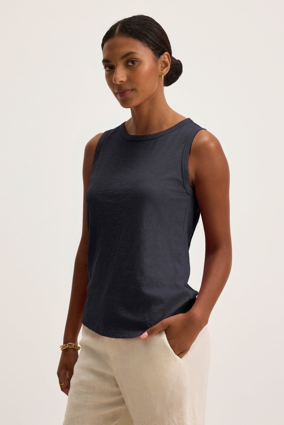 A woman wears the TAURUS TANK TOP by Velvet by Graham & Spencer, featuring a sleeveless, crew neck navy design, paired with light pants as she stands with one hand in her pocket against a plain background.