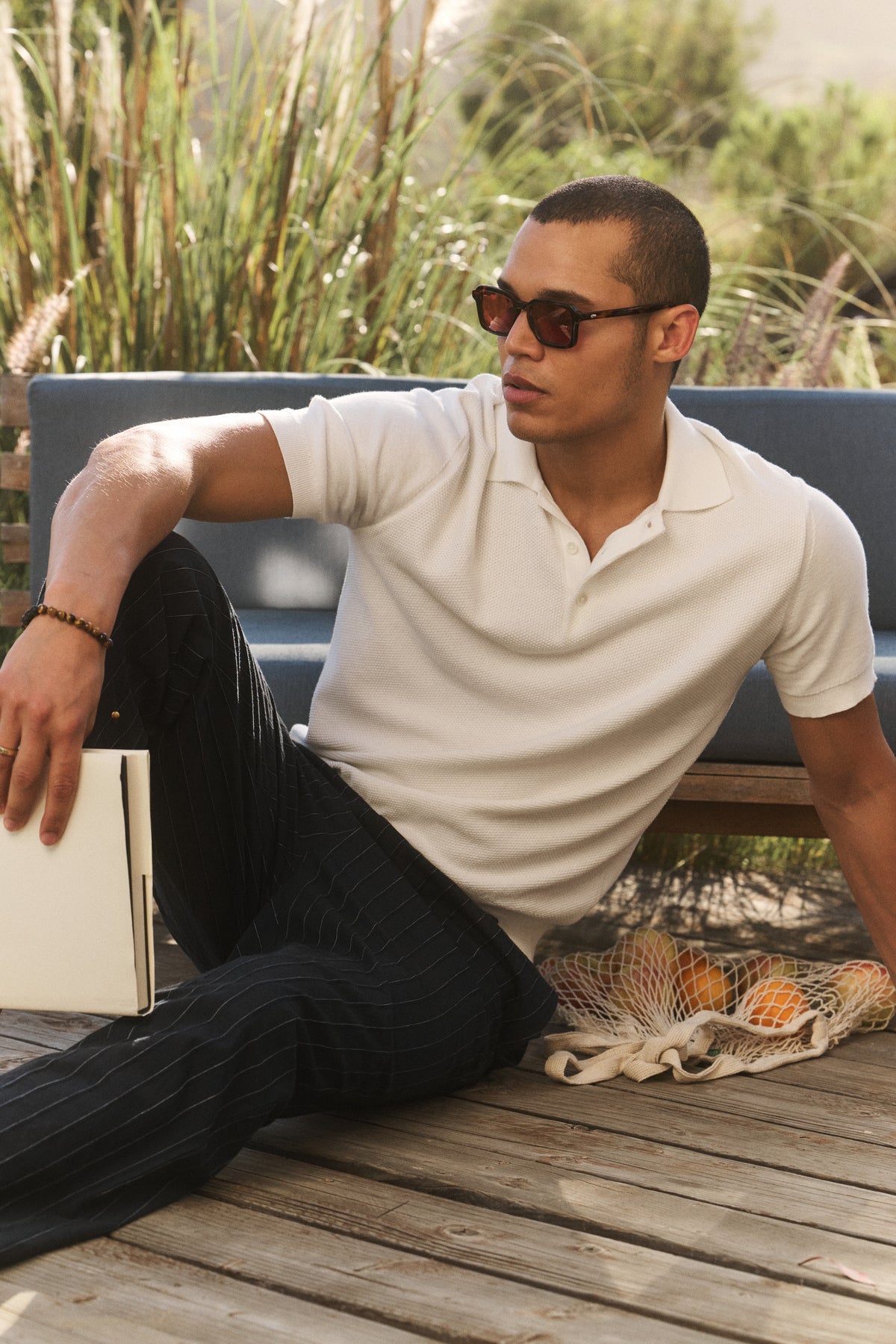 Wearing the DILLON SWEATER POLO by Velvet by Graham & Spencer, a man in sunglasses relaxes on a wooden deck with a closed laptop and a net bag of oranges beside him, enjoying this perfect warm-weather layer.-41617558995137