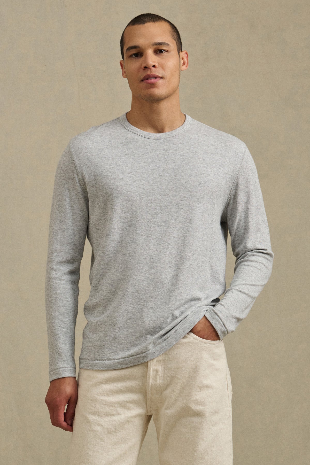 A man wears the COLTON CREW by Velvet by Graham & Spencer—a light gray, long-sleeve crew shirt in soft double-face jersey—with off-white pants, standing against a plain beige background.-41008943169729