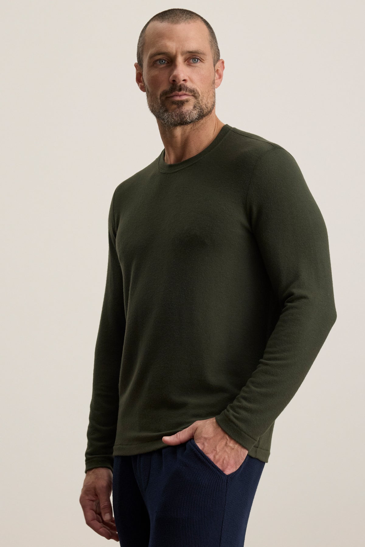 A man with short hair and a beard wears the Velvet by Graham & Spencer BECKER CREW, an olive green fitted crew neck long-sleeve shirt, paired with dark pants—ideal for daily wear—standing against a plain light background, one hand in his pocket.