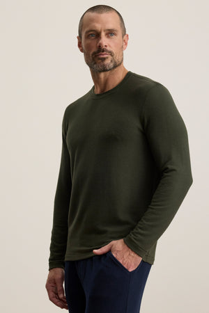 A man with short hair and a beard wears the Velvet by Graham & Spencer BECKER CREW, an olive green fitted crew neck long-sleeve shirt, paired with dark pants—ideal for daily wear—standing against a plain light background, one hand in his pocket.