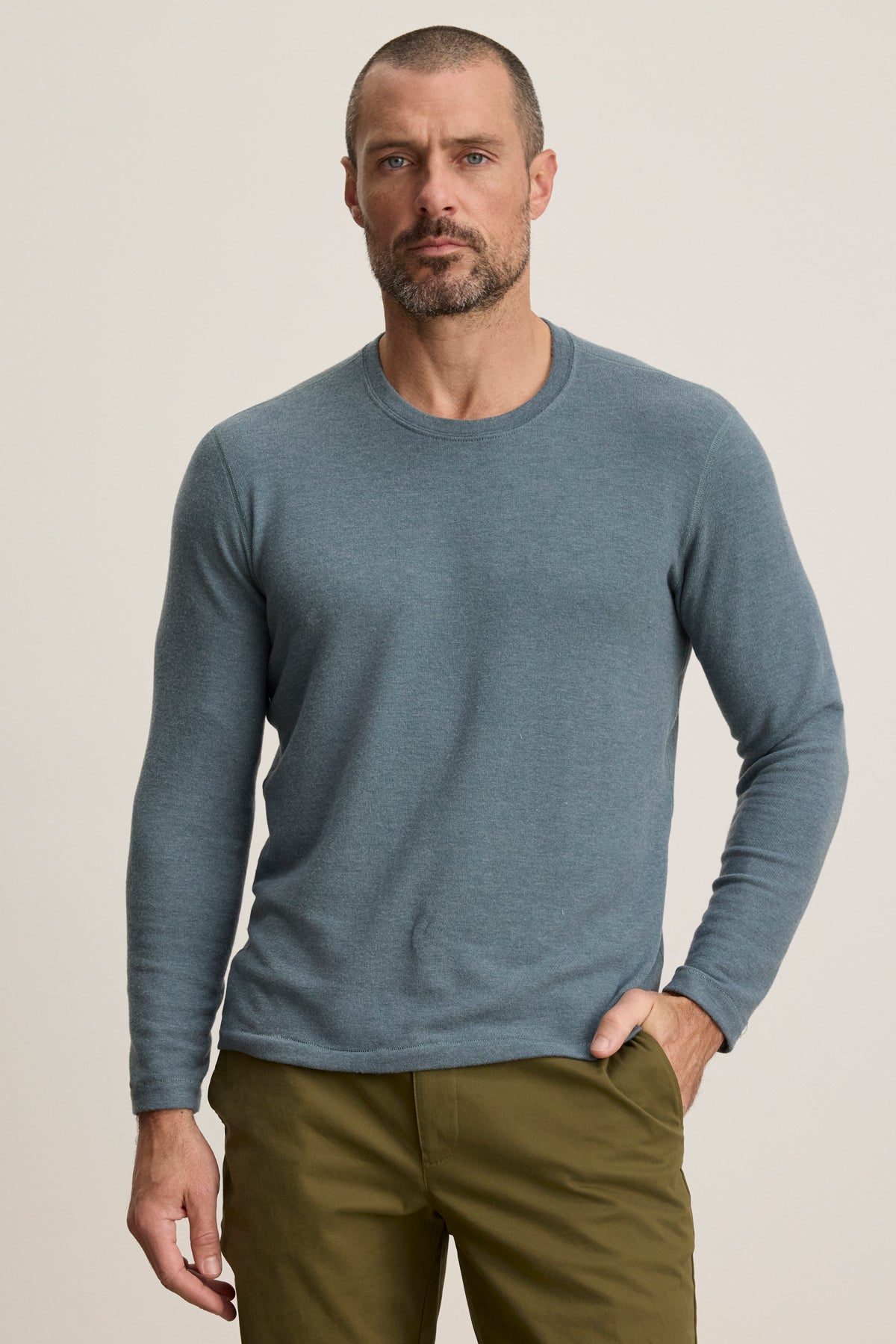 A man with short hair and a beard wears the Velvet by Graham & Spencer BECKER CREW jersey light blue long-sleeve shirt and olive green pants, standing against a plain light background.