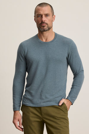 A man with short hair and a beard wears the Velvet by Graham & Spencer BECKER CREW jersey light blue long-sleeve shirt and olive green pants, standing against a plain light background.