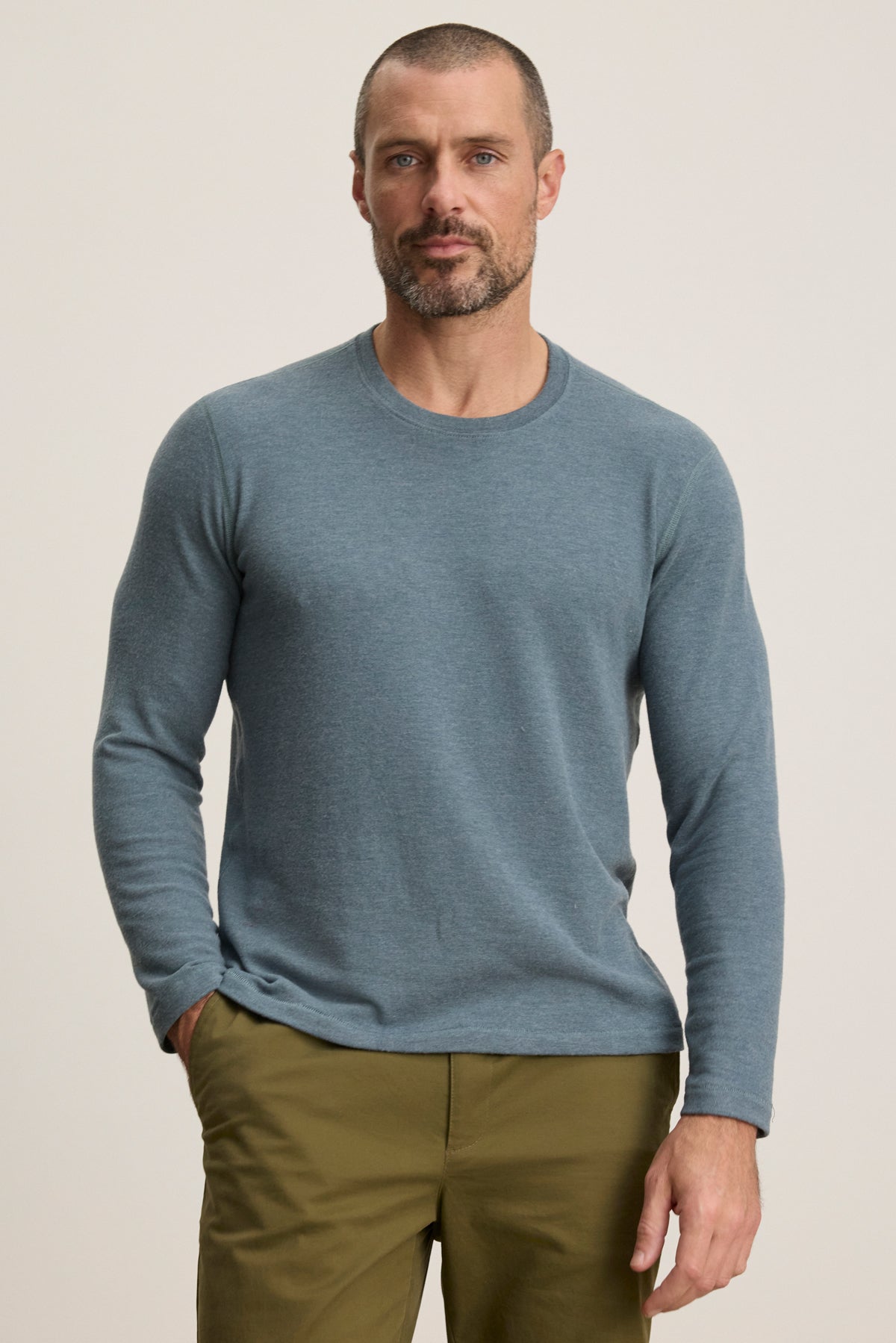 A man with short hair and a beard wears the Velvet by Graham & Spencer BECKER CREW in light blue and olive green pants, standing against a plain light background.