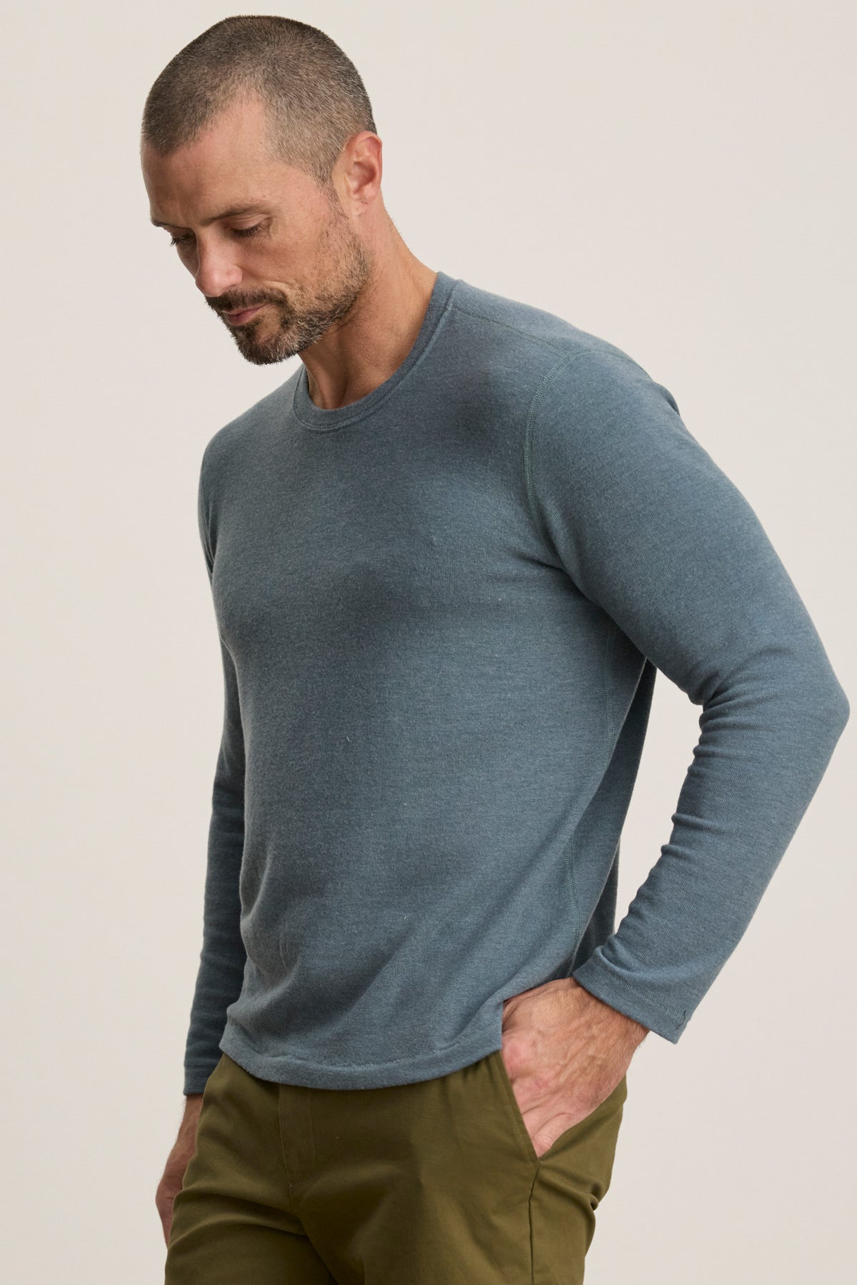 A man with short hair and a beard wears the BECKER CREW blue jersey long-sleeve shirt by Velvet by Graham & Spencer and olive green pants, looking down with one hand in his pocket against a plain background.
