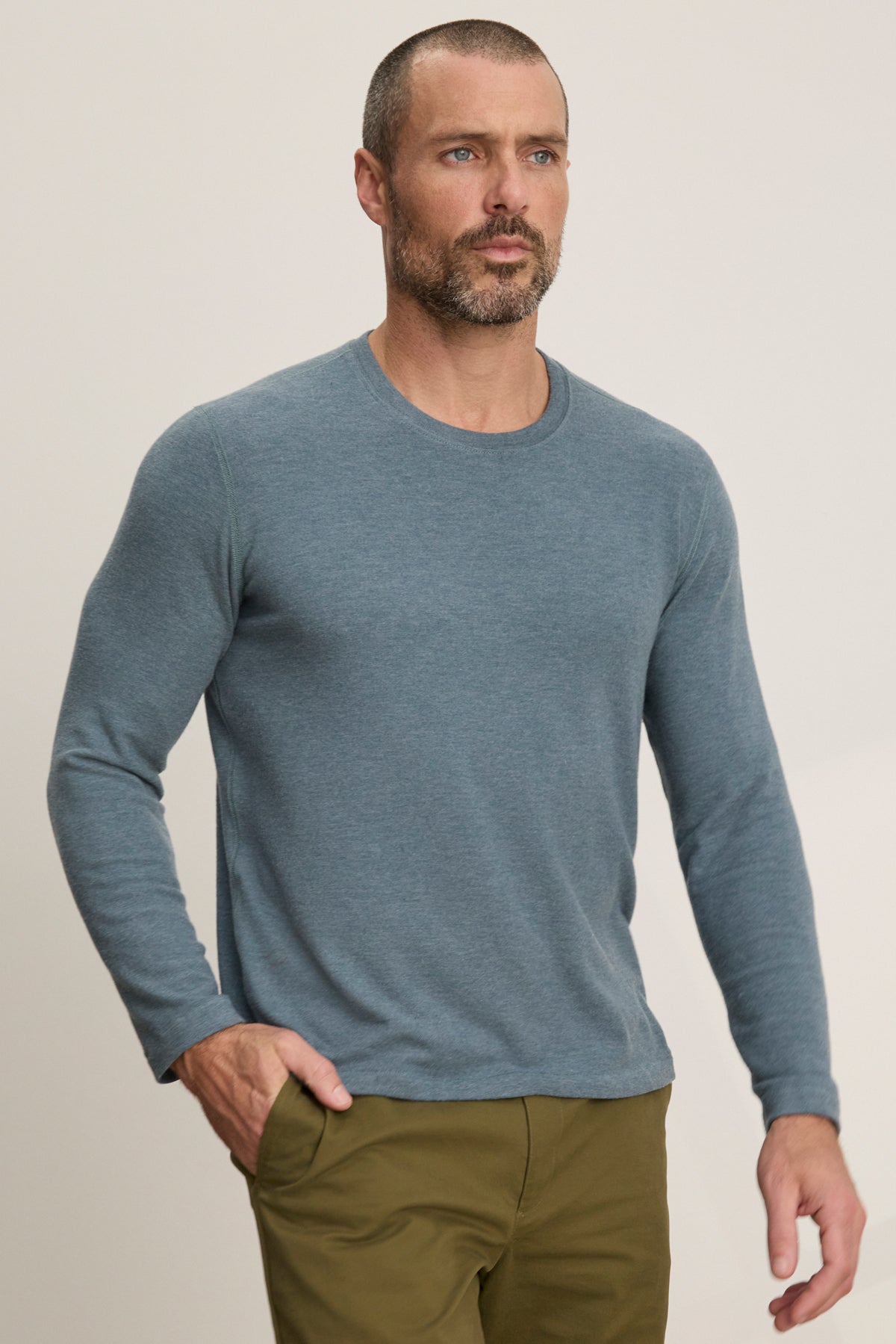 A man with short hair and a beard wears the BECKER CREW by Velvet by Graham & Spencer, a blue fitted crew neck long-sleeve shirt paired with olive green pants, ideal for daily wear, standing against a plain light background.