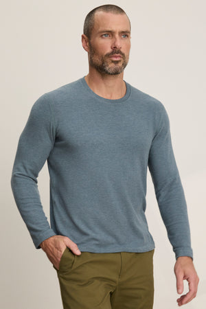 A man with short hair and a beard wears the BECKER CREW by Velvet by Graham & Spencer, a blue fitted crew neck long-sleeve shirt paired with olive green pants, ideal for daily wear, standing against a plain light background.