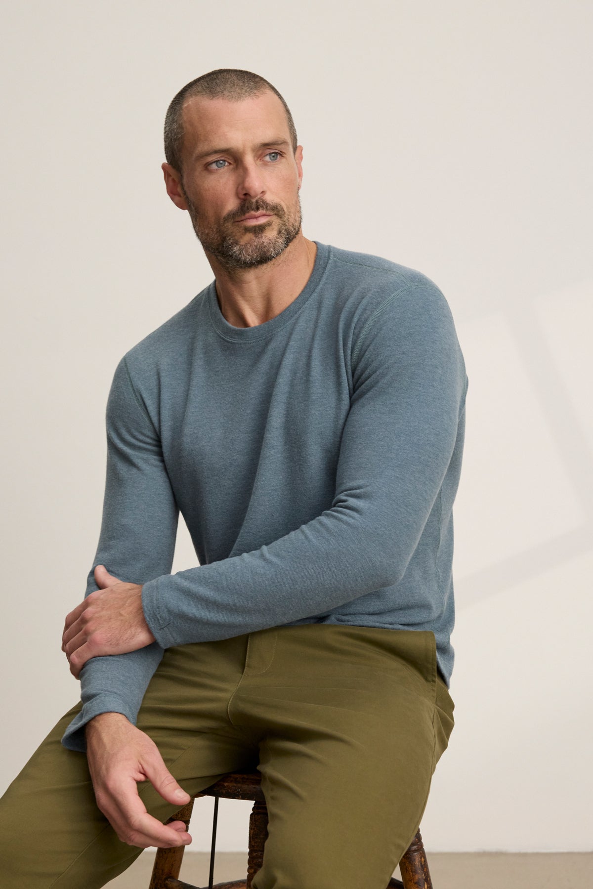 A man with short hair and a beard, dressed in Velvet by Graham & Spencer's BECKER CREW blue long-sleeve shirt and olive pants, sits on a wooden stool—a simple, stylish choice for effortless daily wear.