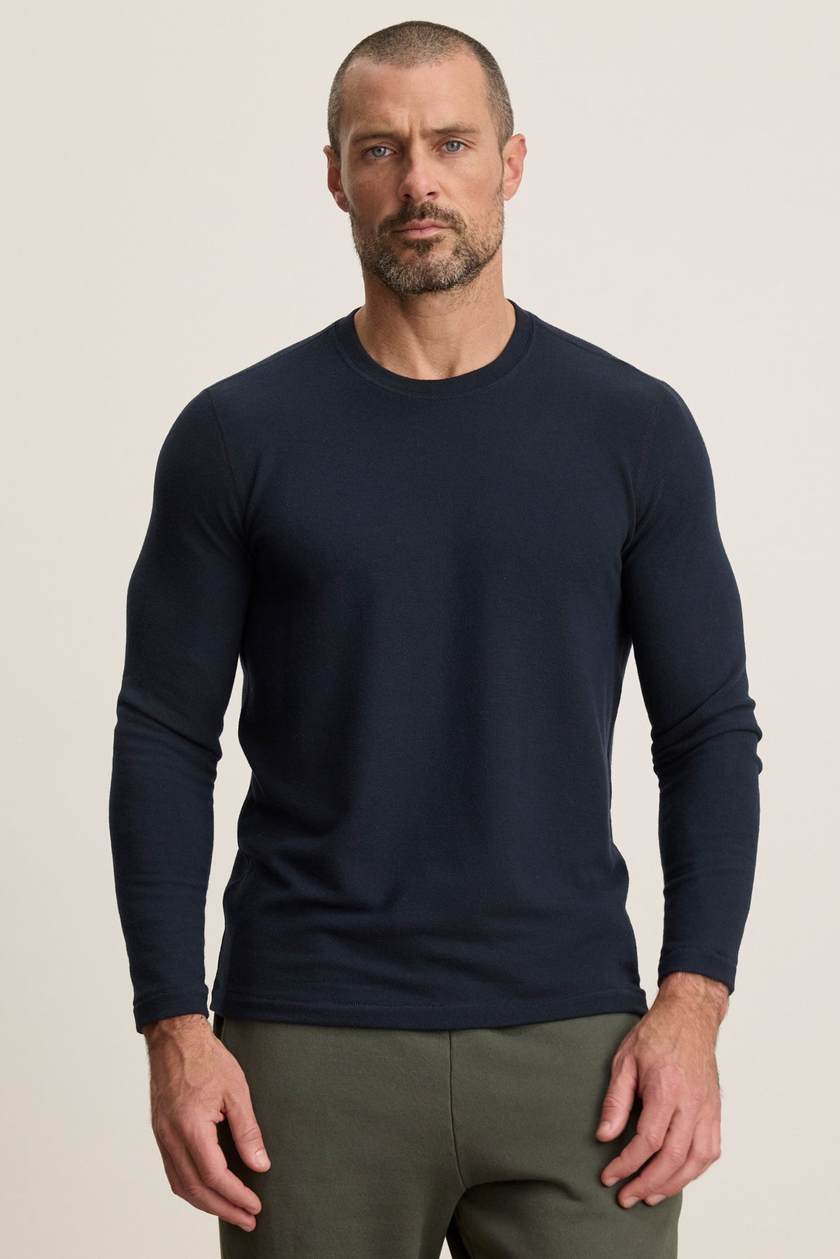 A man with short hair and a beard wears the BECKER CREW by Velvet by Graham & Spencer, a fitted navy jersey shirt for daily comfort, paired with olive green pants, standing against a plain light background.