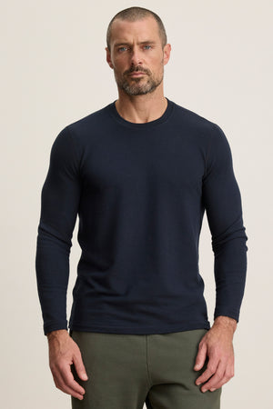 A man with short hair and a beard wears the BECKER CREW by Velvet by Graham & Spencer, a fitted navy jersey shirt for daily comfort, paired with olive green pants, standing against a plain light background.