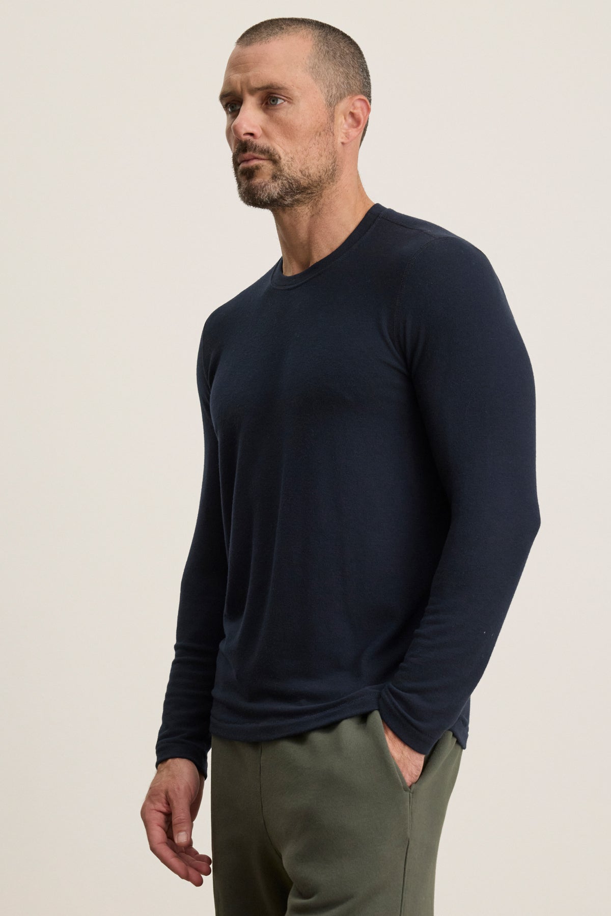 A man with short hair and a beard wears the BECKER CREW, a fitted navy long-sleeve by Velvet by Graham & Spencer, paired with olive pants—perfect for daily wear—standing with one hand in his pocket against a plain light background.