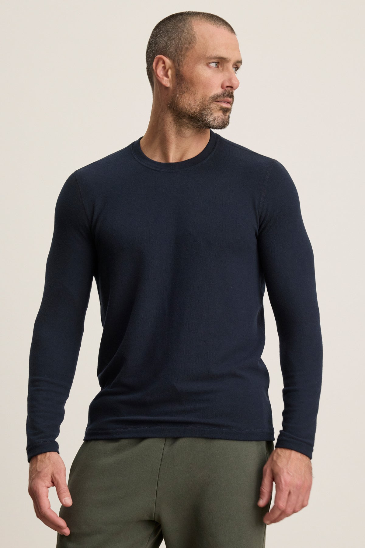 A man with a close-cropped beard and short hair wears the Velvet by Graham & Spencer BECKER CREW, a fitted navy blue long-sleeve jersey, with olive green pants against a beige background—perfect for stylish daily wear.