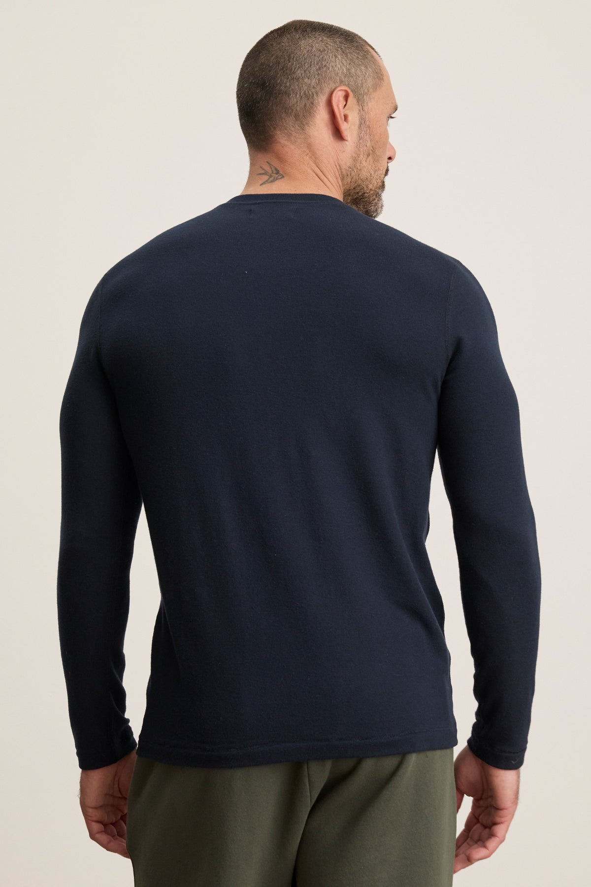 A man with short hair and a neck tattoo is seen from the back, wearing the Velvet by Graham & Spencer BECKER CREW in navy blue and olive green pants—an ideal daily wear look against a plain background.