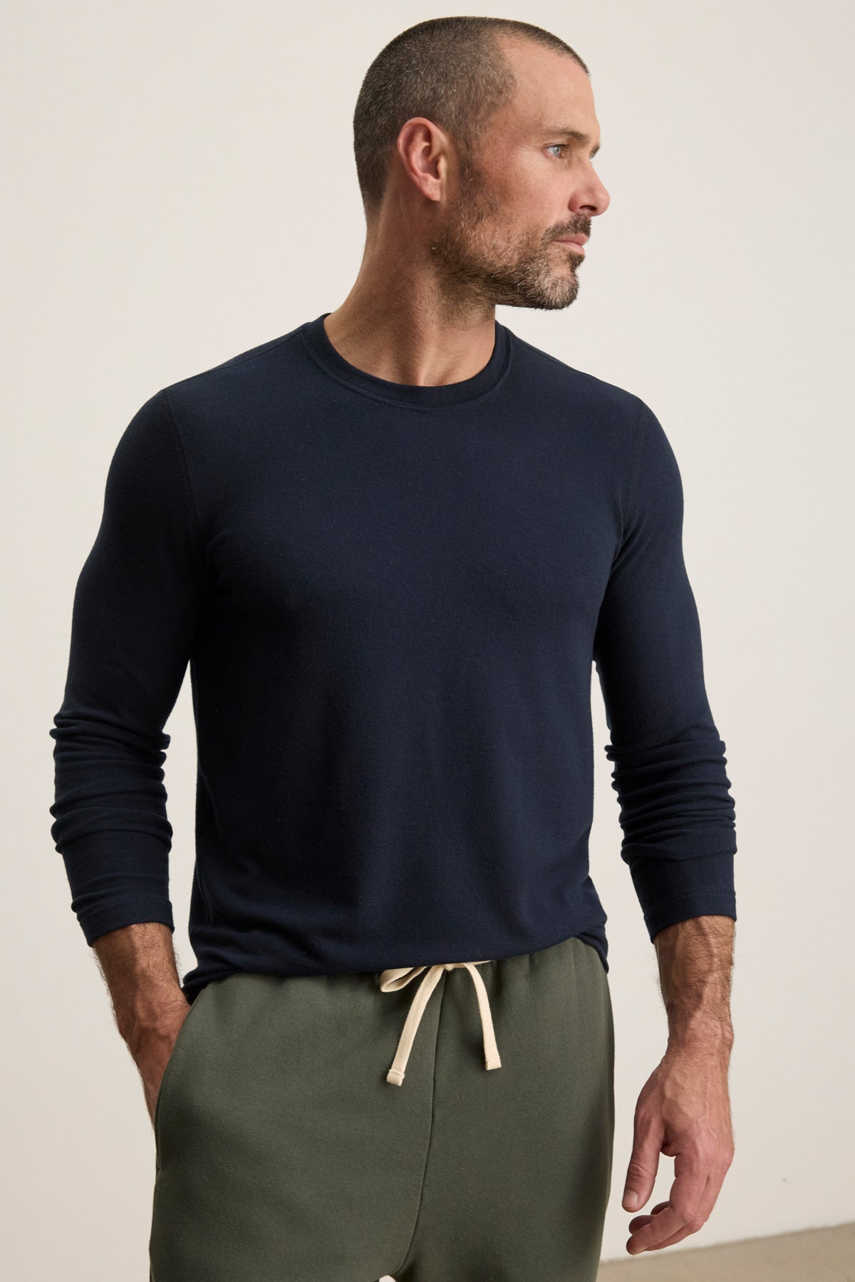 A man with a short beard models the BECKER CREW by Velvet by Graham & Spencer—a fitted navy long-sleeve shirt—paired with olive drawstring pants, standing with hands in pockets and looking to his right; ideal for comfortable daily wear.