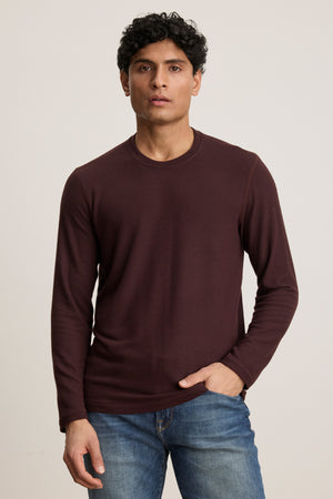 A man with short curly hair wears the Velvet by Graham & Spencer BECKER CREW, a fitted maroon long-sleeve shirt, paired with blue jeans, standing against a plain light background.