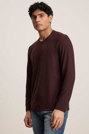 A man wearing the BECKER CREW sweater in burgundy by Velvet by Graham & Spencer and blue jeans stands against a plain light background, looking at the camera.