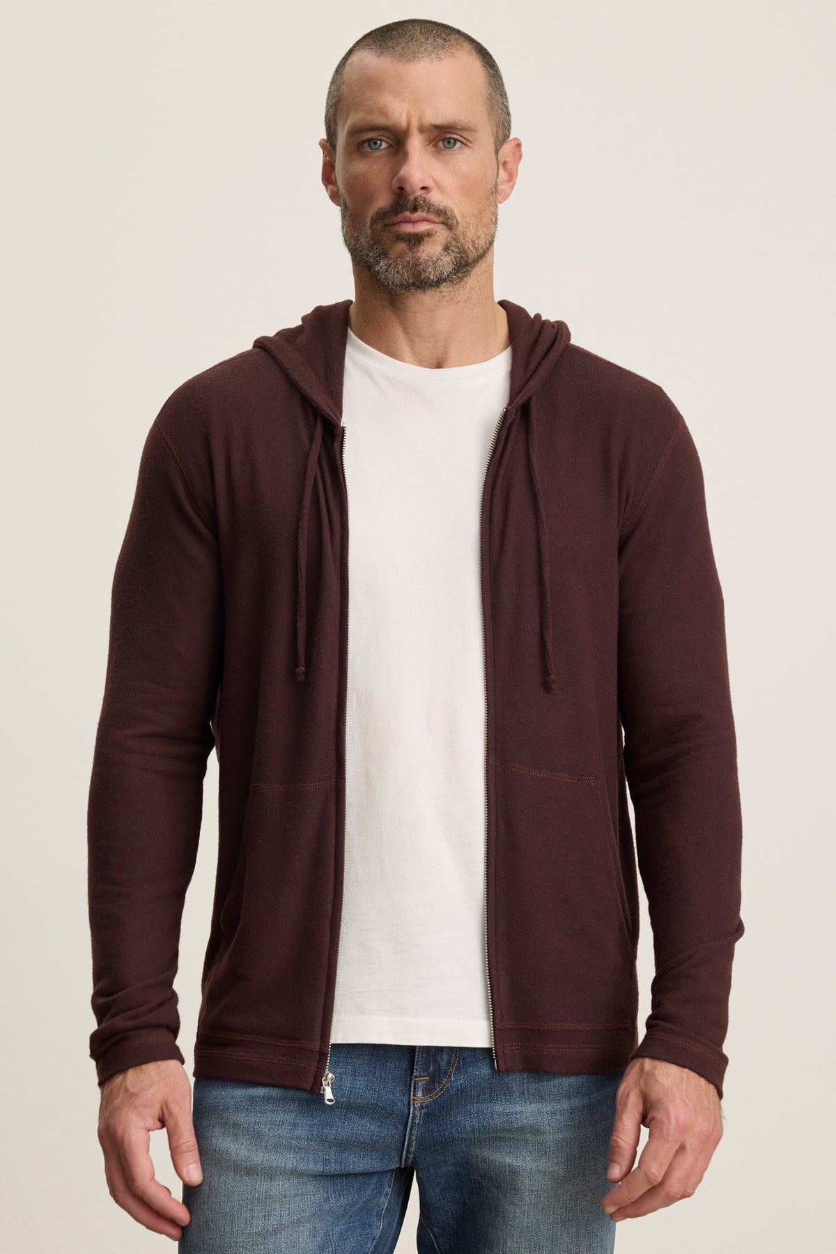 A man stands against a plain background, wearing a white t-shirt, blue jeans, and the LOUDON HOODIE by Velvet by Graham & Spencer—a relaxed fit maroon full-zip hoodie crafted from triblend cozy jersey.